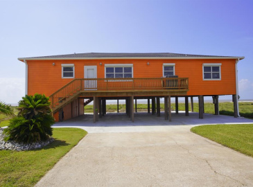 This is an elevated, single-story, orange home with a wooden deck and ample under-house parking. The property features a landscaped front yard and offers a clear view of the horizon.