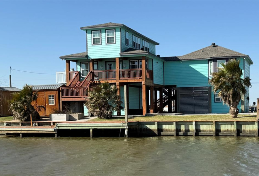 This expansive waterfront home features a vibrant turquoise exterior with a raised two-story design, offering picturesque views and ample outdoor living space. The property includes a water view deck, lush palm trees, and direct access to the water, perfect for fishing, entertaining and relaxing.