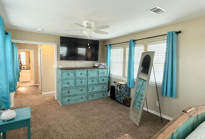 This spacious bedroom features large windows for natural light when desired, and a ceiling fan. It includes a turquoise dresser, a wall-mounted TV. The room connects to a bathroom, enhancing convenience and comfort.