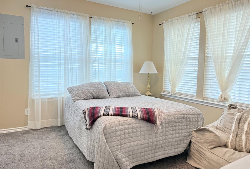 This cozy bedroom features a comfortable bed with a quilted cover, ample natural light from multiple windows with sheer curtains, and soft carpeting. A small side table with a lamp adds functionality.