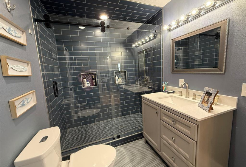 Modern bathroom/laundry room features a sleek glass-enclosed shower with dark subway tiles, a stylish vanity with ample storage, and contemporary lighting. With stacked washer and dryer it has a clean, sophisticated design ideal for a comfortable and functional home space.