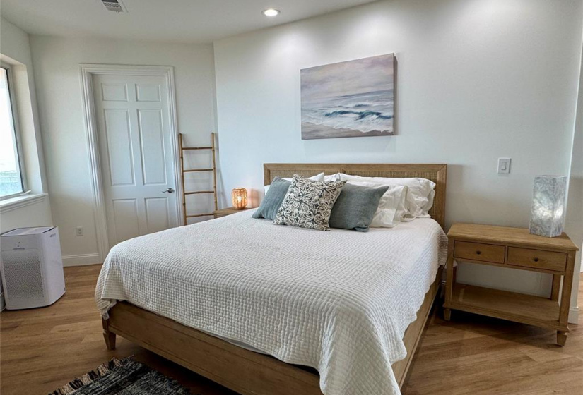This cozy bedroom features a large bed with a wooden frame, complemented by a side table and stylish decor. The room is well-lit with recessed lighting and a window allowing natural light. A serene painting adds a calming touch to the modern, minimalist design.