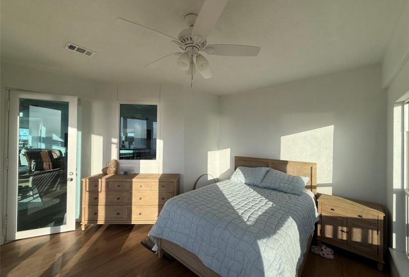 Bright bedroom with natural light, featuring a ceiling fan, hardwood floors, and light wood furniture. There's a cozy bed, dresser, and nightstand, with a door leading to a patio or balcony area.