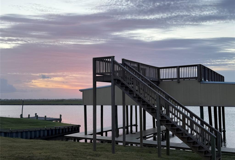 This photo showcases a serene waterfront view at sunset, featuring a wooden dock with stairs leading to an elevated deck. The calm water and expansive sky create a tranquil outdoor setting, perfect for relaxation or entertaining.